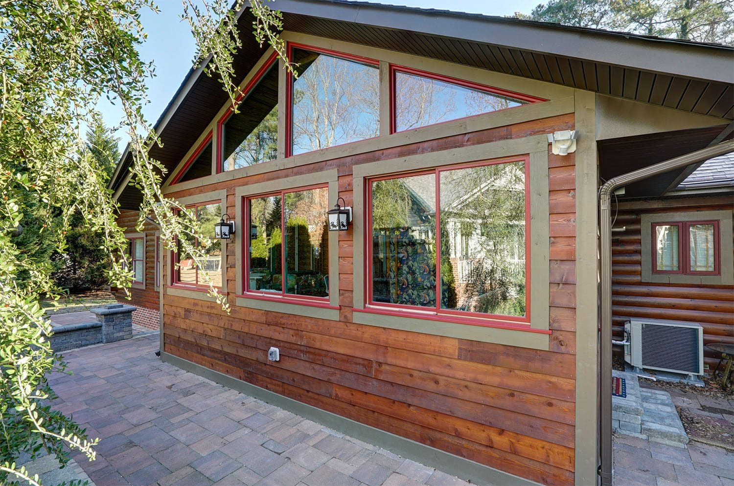 Rustic cabin-style sunroom with stained wood siding, adorned with red trim around glass windows. A stone hardscape patio, designed and built by Deck Creations, extends from the sunroom, surrounded by nature and greenery. Bathed in sunlight, this cozy space harmonizes with its natural surroundings.
