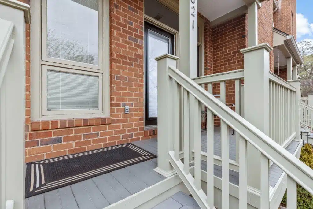 A tastefully designed covered porch and staircase, constructed by Deck Creations for a commercial property. The stairs gracefully descend to a small hardscape area featuring inviting patio chairs and well-manicured shrubs.