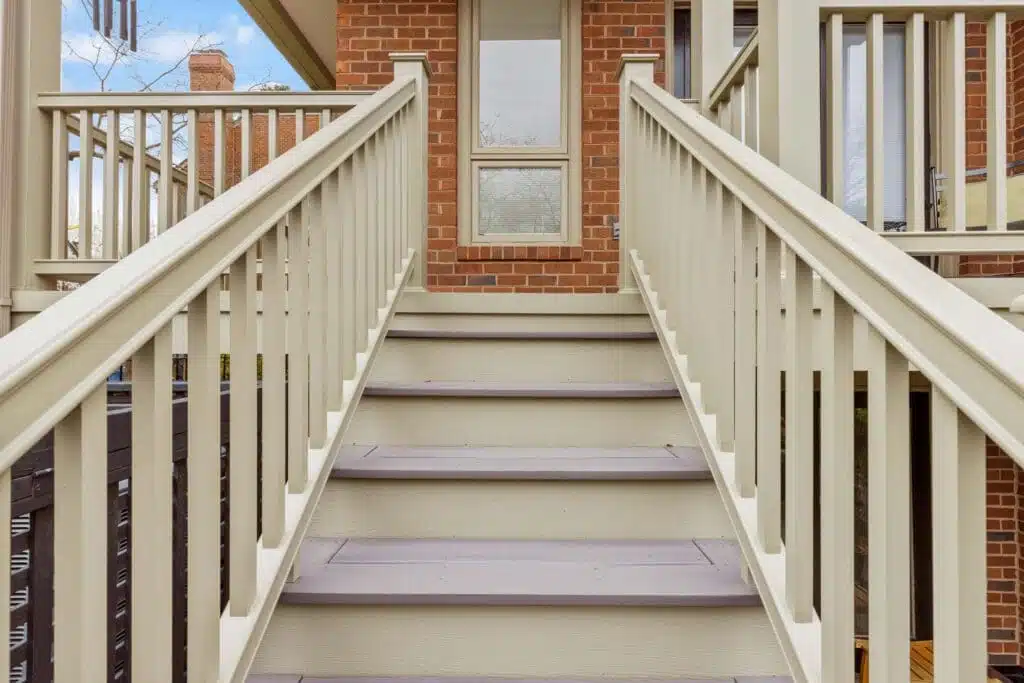 A tastefully designed covered porch and staircase, constructed by Deck Creations for a commercial property. The stairs gracefully descend to a small hardscape area featuring inviting patio chairs and well-manicured shrubs.