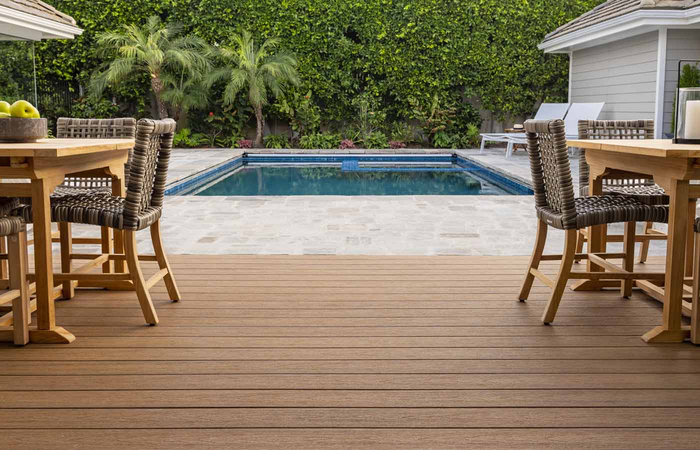 A spacious wooden deck with two tables sits gracefully near a shimmering swimming pool, enveloped by lush greenery and accented by a charming white house.