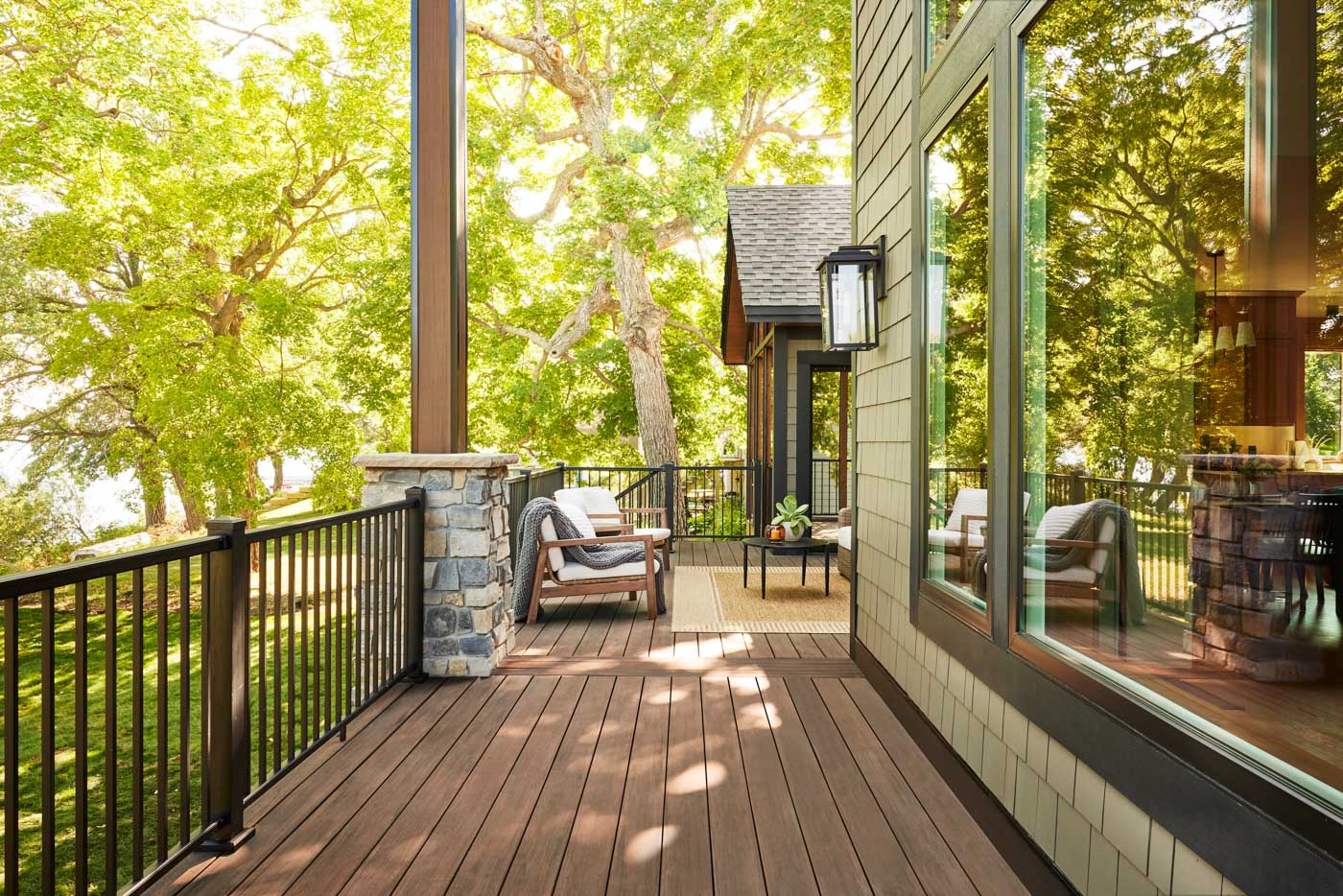 The wooden deck, adorned with chairs and a small table, is framed by a stone pillar railing. Large glass windows reflect the surrounding green trees as sunlight filters through the lush canopy, creating an idyllic outdoor retreat.