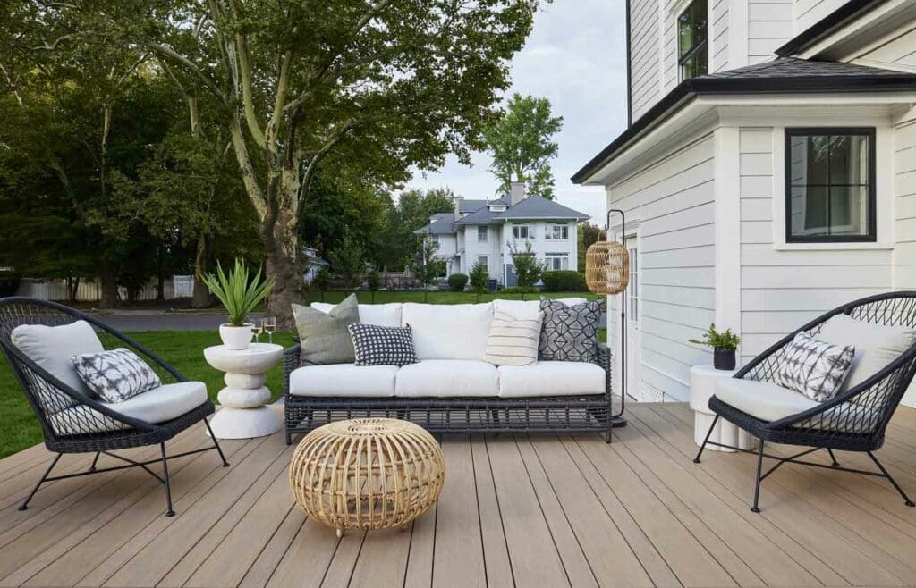 A modern outdoor patio features a white cushioned sofa, two black chairs, and a round wicker table amid lush potted plants. Set on a spacious wooden deck, this inviting space is framed by surrounding trees and nearby houses.