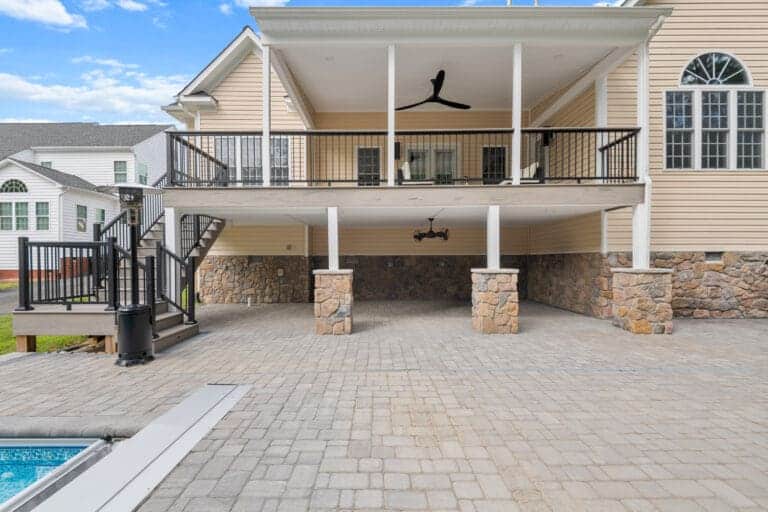 Elevated patio with ceiling fan above stone-supported carport, adjacent to a pool. Brick driveway and exterior stairs visible, leading to the upper level of a beige house.