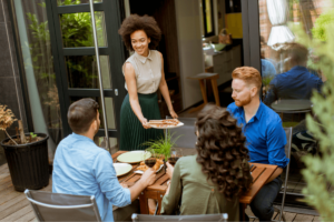A woman serves food to three people gathered around an outdoor table on a patio, embodying the essence of outdoor living with its charming potted plants.