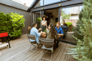 Five people savoring a meal and drinks on a wooden deck outside a modern building, embracing the essence of outdoor living, with lush greenery and a barbecue grill adding to the serene ambiance.