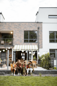 Four people sitting on a step, enjoying drinks, embrace the essence of outdoor living in front of a modern house adorned with brick accents and a spacious patio.