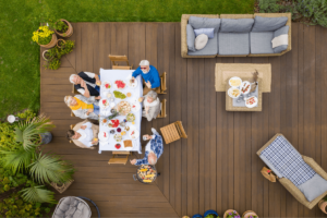 Aerial view of a group of people enjoying outdoor living, sitting around a dining table on a wooden deck adorned with various food items, surrounded by cozy furniture and lush greenery.