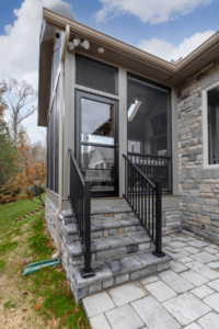 The image shows a stone patio with steps leading to a screened porch, complemented by sleek black railings. A glass door opens to the space, while the charming siding of the house stands in contrast against a backdrop of trees and a cloudy sky.