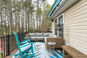 A cozy outdoor deck features a wicker sectional sofa, blue rocking chairs, a small round table, and a leaf-patterned rug, all surrounded by trees.