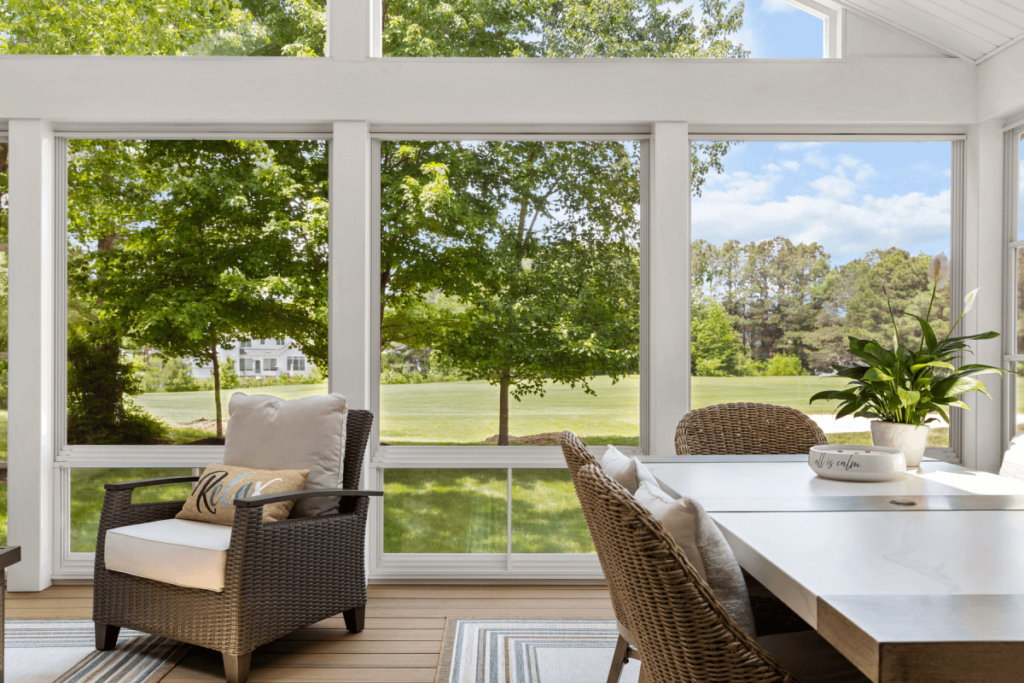 The bright sunroom, framed by elegant siding, boasts large windows overlooking a green lawn and features wicker seating, a white table, and a potted plant.