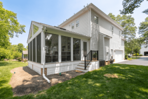 The charming white two-story house features elegant siding and a screened porch, all surrounded by a lush green lawn and trees under a clear blue sky.