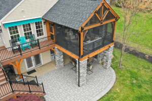 Aerial view of a house with stylish siding, showcasing a covered patio featuring stone pillars, screened porch, and outdoor seating on a paved area, all nestled amidst lush green grass and trees.