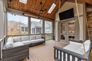 A cozy enclosed patio with wicker sofas, a hanging bench, and a ceiling fan seamlessly integrates wood paneling and siding. Skylights brighten the space, which also features a wall-mounted TV, offering serene views of the backyard.