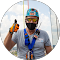 A person in safety gear, including a helmet, mask, gloves, and harness for siding work, gives a thumbs-up gesture while holding a radio device. Clouds are visible in the background.