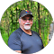 A man with a gray beard and glasses, dressed in a black cap and shirt, enjoys the essence of outdoor living amid towering trees.