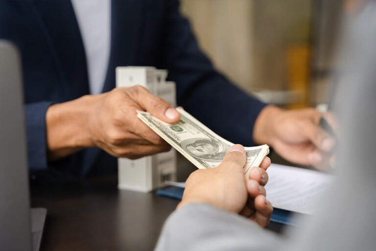 Amidst the vibrant ambiance of outdoor living, a person hands over a stack of U.S. dollar bills to another across a picnic table, capturing the essence of a breezy al fresco transaction.