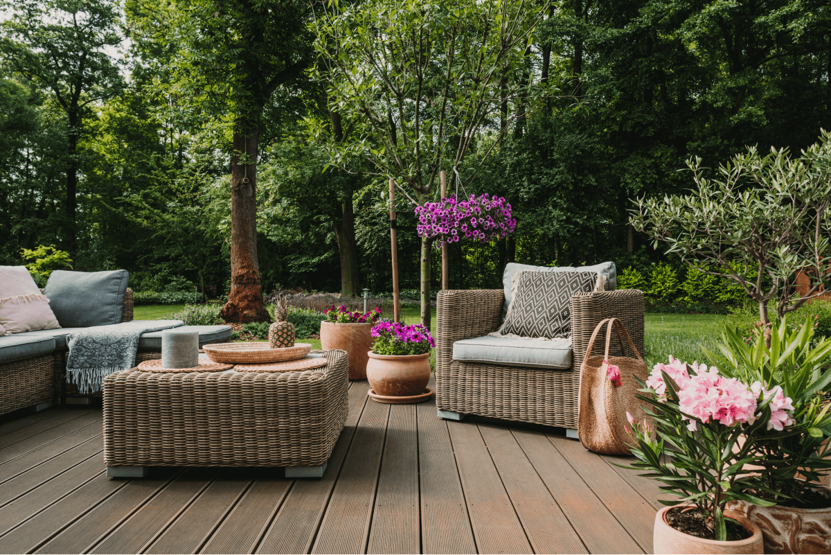 A cozy outdoor patio designed for perfect outdoor living, featuring wicker furniture, cushions, and potted plants with pink and purple flowers set against a backdrop of lush greenery.