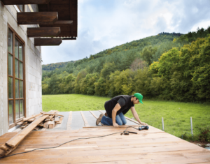 A person wearing a green cap is kneeling and working on a wooden deck with a tool, enhancing their outdoor living space, surrounded by a scenic green landscape.