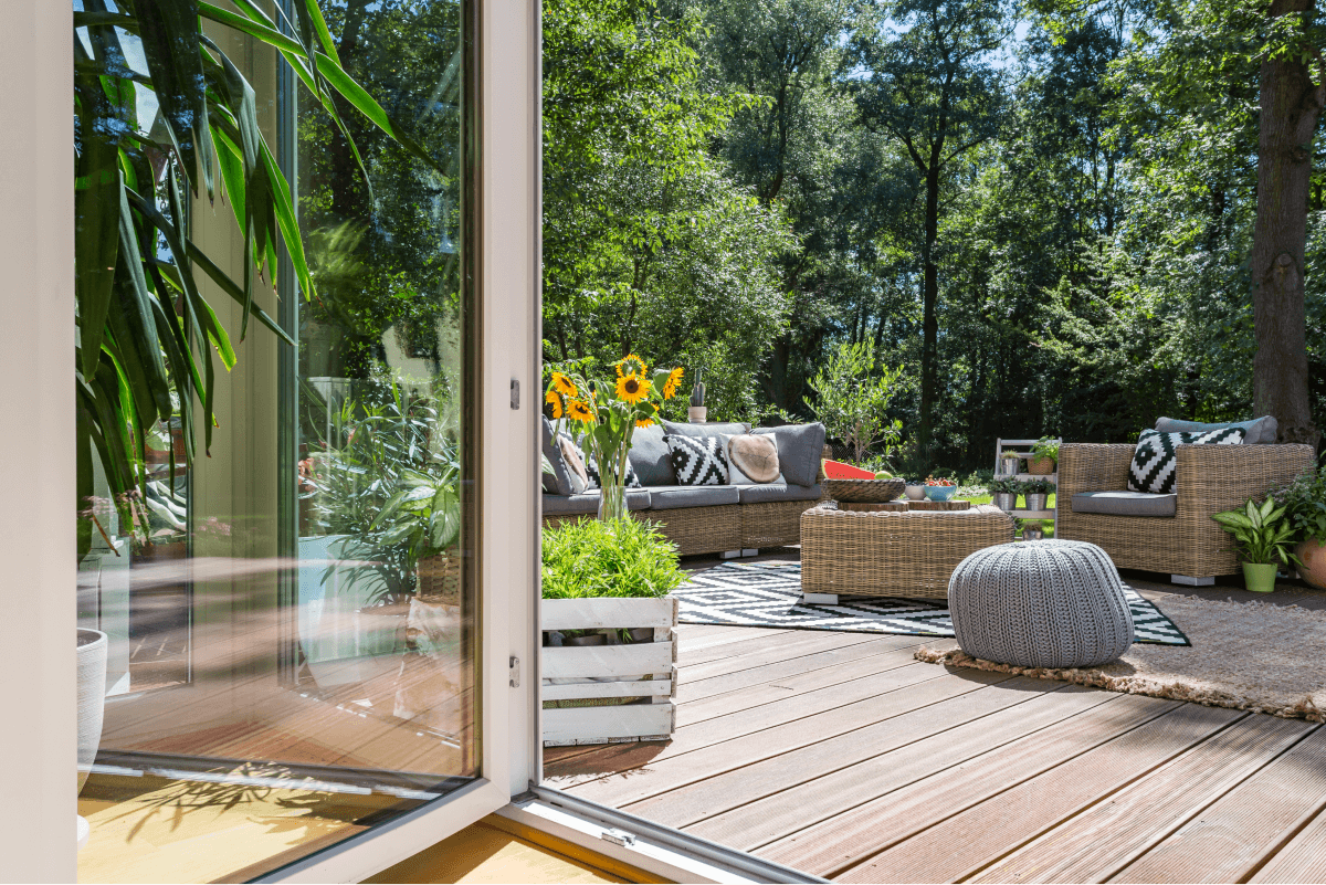 Open patio door leading to a deck designed for outdoor living, complete with wicker furniture, sunflowers, and lush greenery. A serene forested area provides a calming backdrop.