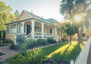 A charming yellow house with a porch perfect for outdoor living, surrounded by a lush garden, and the sun shining through the trees. An American flag flutters near the entrance, while a white picket fence lines the front yard.