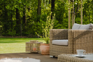 A wicker chair with cushions, a potted plant, and a lantern create the perfect outdoor living space on a deck, complete with a cup for leisurely sipping while overlooking the grassy lawn and trees.