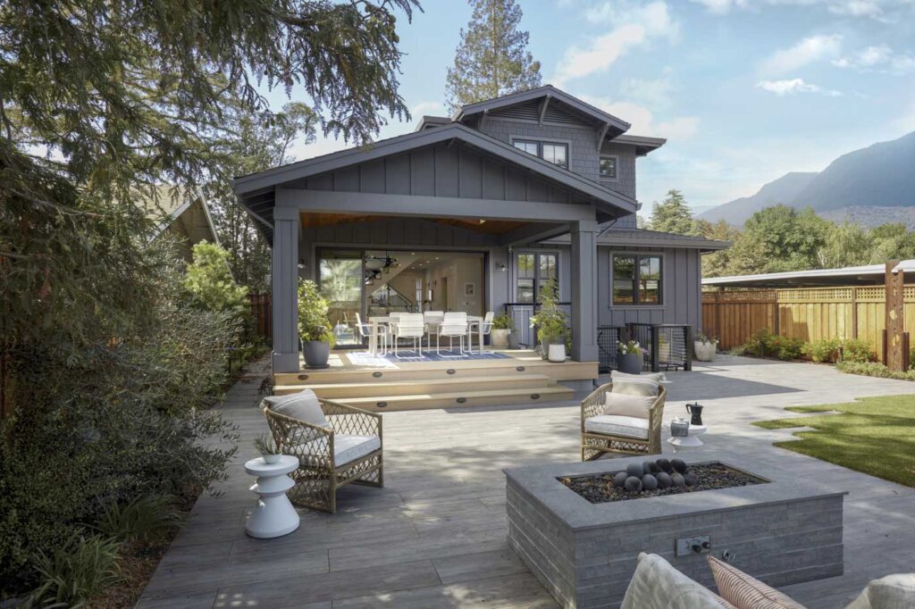 Modern two-story house with a gray exterior, nestled among trees and mountains. A spacious deck with wicker furniture and a firepit transitions to a lawn and garden area under the blue sky.