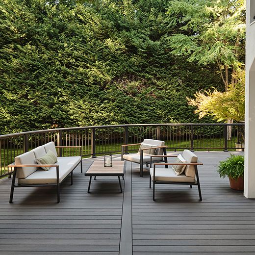 The outdoor patio, featuring a spacious gray deck, offers a cozy seating area with a sofa, two chairs, and a coffee table. It's beautifully enclosed by metal railings and surrounded by dense greenery and towering trees in the background.