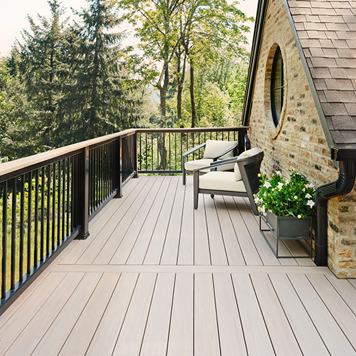 A charming wooden deck with two chairs and a small table extends from a brick house featuring a round window. This cozy haven is surrounded by lush trees and greenery, perfect for enjoying nature's serenity.