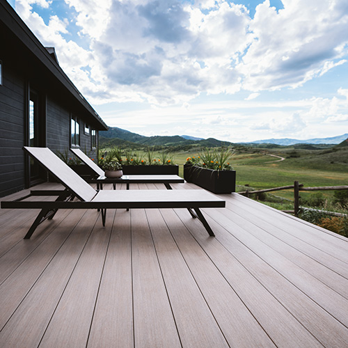 A sleek, contemporary deck with two lounge chairs offers a serene view of the vast, green landscape beneath a partly cloudy sky.