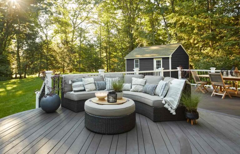 The cozy outdoor deck features a wicker sectional sofa and round ottoman, nestled amidst the trees. A small black shed graces the background, while a dining table sits to the right, perfect for alfresco meals.