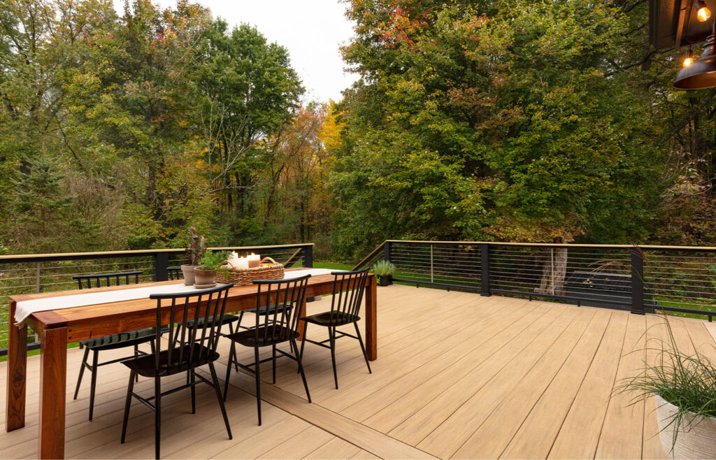 The inviting outdoor deck, featuring a wooden dining table and six chairs, is enveloped by tall trees bearing green and autumnal leaves. A white table runner graces the deck's centerpiece alongside a small decorative plant.