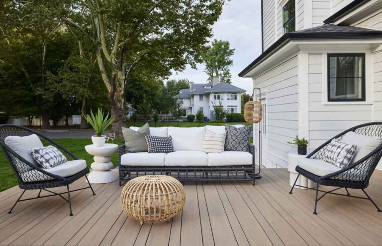 The outdoor patio features a white sofa, two black wicker chairs, and a rattan coffee table on a spacious deck, complemented by a potted plant. Trees and houses provide a charming backdrop.