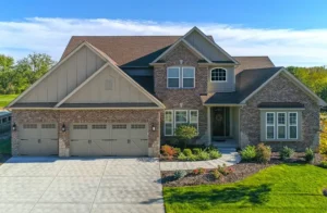 A charming two-story brick house with elegant siding features a three-car garage, front garden, and spacious driveway. The pitched roof stands proudly against a clear sky dotted with a few clouds.