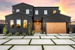 Modern black house with sleek siding, large windows, wooden garage door, and landscaped front yard under a colorful sunset sky.