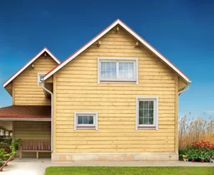 A charming wooden house with a red roof and white-framed windows features elegant siding. It boasts a cozy side porch, set against a backdrop of blue sky and lush plants surrounding the property.