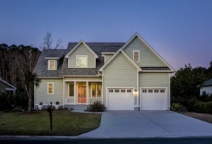 A two-story suburban house with siding, a double garage, lit windows, and a manicured lawn is shown at dusk under a clear sky.
