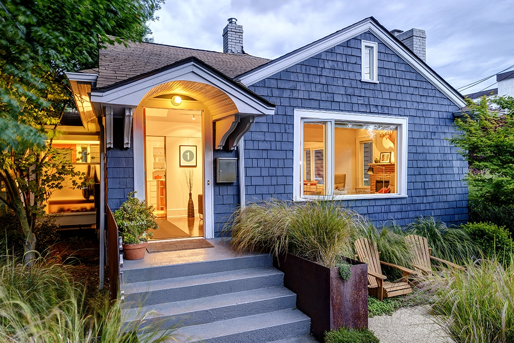 A blue, single-story house features charming siding and a welcoming porch with steps leading up. Large windows reveal cozy interior lighting, while the landscaping includes lush grasses and a path adorned with two wooden chairs in front.