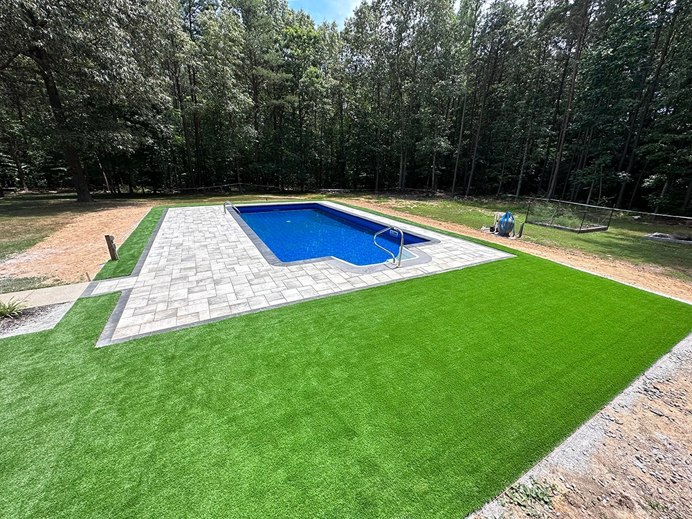 This rectangular swimming pool with shimmering blue water is the centerpiece of an outdoor living oasis, surrounded by a stone patio and lush artificial grass, all set against a backdrop of dense trees.