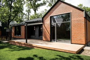 Single-story brick house with large windows and a wooden deck, complemented by sleek siding, surrounded by a grassy lawn and trees.