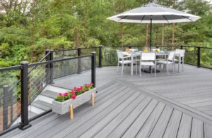 A sleek, modern deck features gray flooring and a glass railing. The dining set, shaded by an umbrella, is surrounded by comfortable chairs. Pink flowers bloom in a box at the edge of this inviting space, with trees providing a serene background to the stylish deck ambiance.