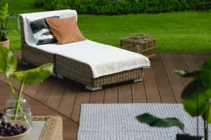 A sleek outdoor wicker chaise lounge with a white cushion and two pillows graces the wooden deck. Surrounded by lush greenery, it pairs perfectly with a matching small wicker side table and a gray patterned rug, creating an inviting retreat on your deck.