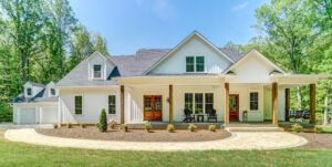 A modern white house with a TImberTech Deck, front porch, wood columns, dormer windows, landscaped yard, and a detached garage, surrounded by tall trees under a clear blue sky.
