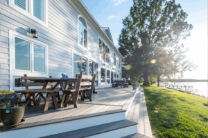 Modern lakeside house with large windows and a spacious wooden deck for outdoor living, featuring comfortable seating, surrounded by green grass and trees, with sunlight reflecting off the water. Ideal for exterior renovation inspiration.