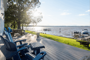 Experience outdoor living on a wooden deck with blue chairs overlooking a calm lake, grassy yard, and boat docks—all perfect for relaxing or planning your next exterior renovation on a sunny day with clear skies.