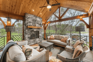 Experience elevated outdoor living on a covered patio featuring a stone fireplace, ceiling fan, two sofas, armchairs, and woven lamps—all beautifully enhanced by an exterior renovation that frames serene views of trees and grass through open railings.