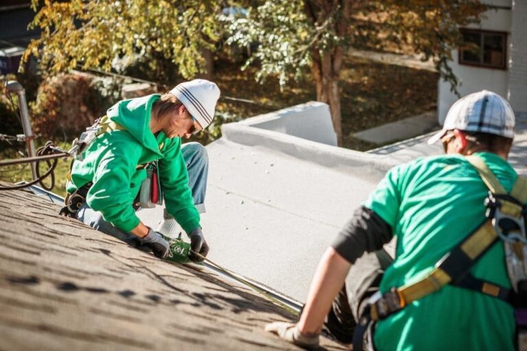 Two workers in green shirts and safety gear install or repair roofing shingles on a sloped roof during daylight.