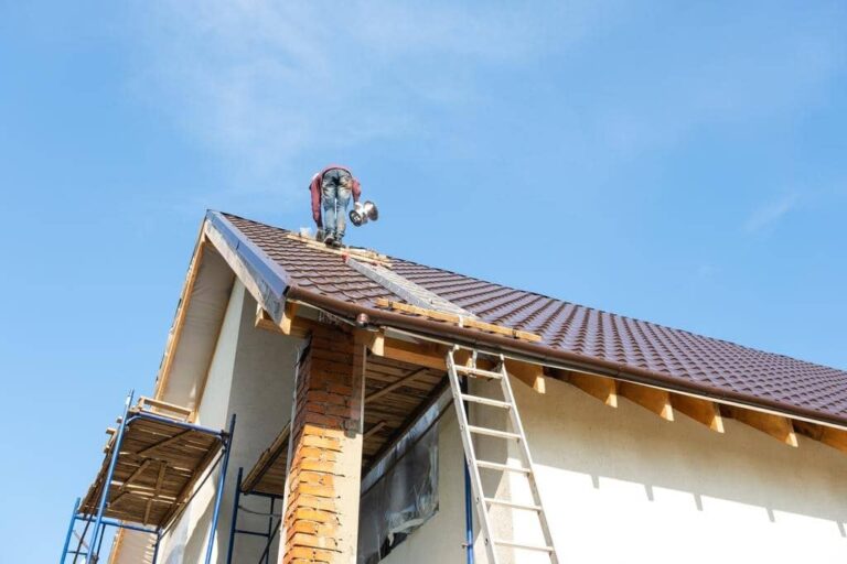 A person stands on the roof of a house under construction, holding tools, with scaffolding and a ladder positioned against the building.
