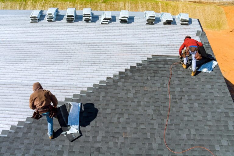 Two workers install dark roof shingles on a sloped roof, with tools and materials scattered nearby and lighter shingles stacked at the top edge.