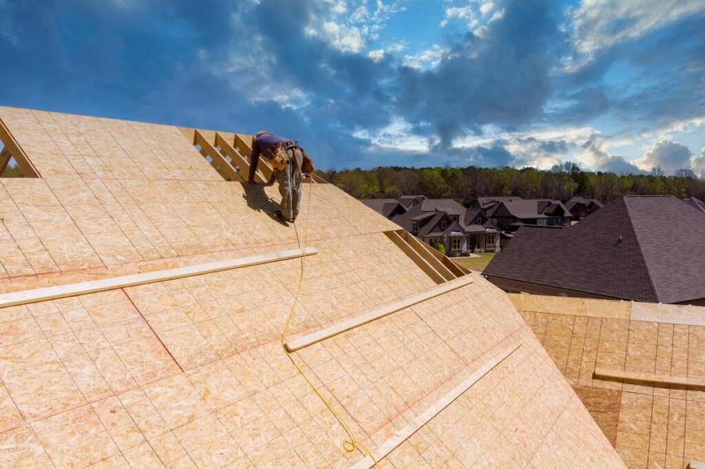 A construction worker installs wooden panels on a sloped roof of a house under construction, with rooftops and trees visible in the background.