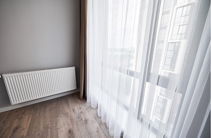 A bright room corner with a wooden floor, a white radiator on the wall, and sheer white curtains covering large windows.